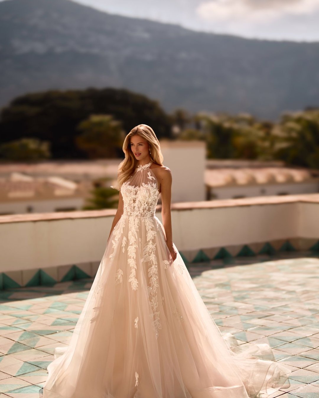 Iltana to romantyczna suknia ślubna z dekoltem halter, wyciętymi plecami i dekoracyjnymi aplikacjami w kształcie liści. Zachwyca szeroką, tiulową spódnicą, delikatnym połyskiem i subtelnym trenem.