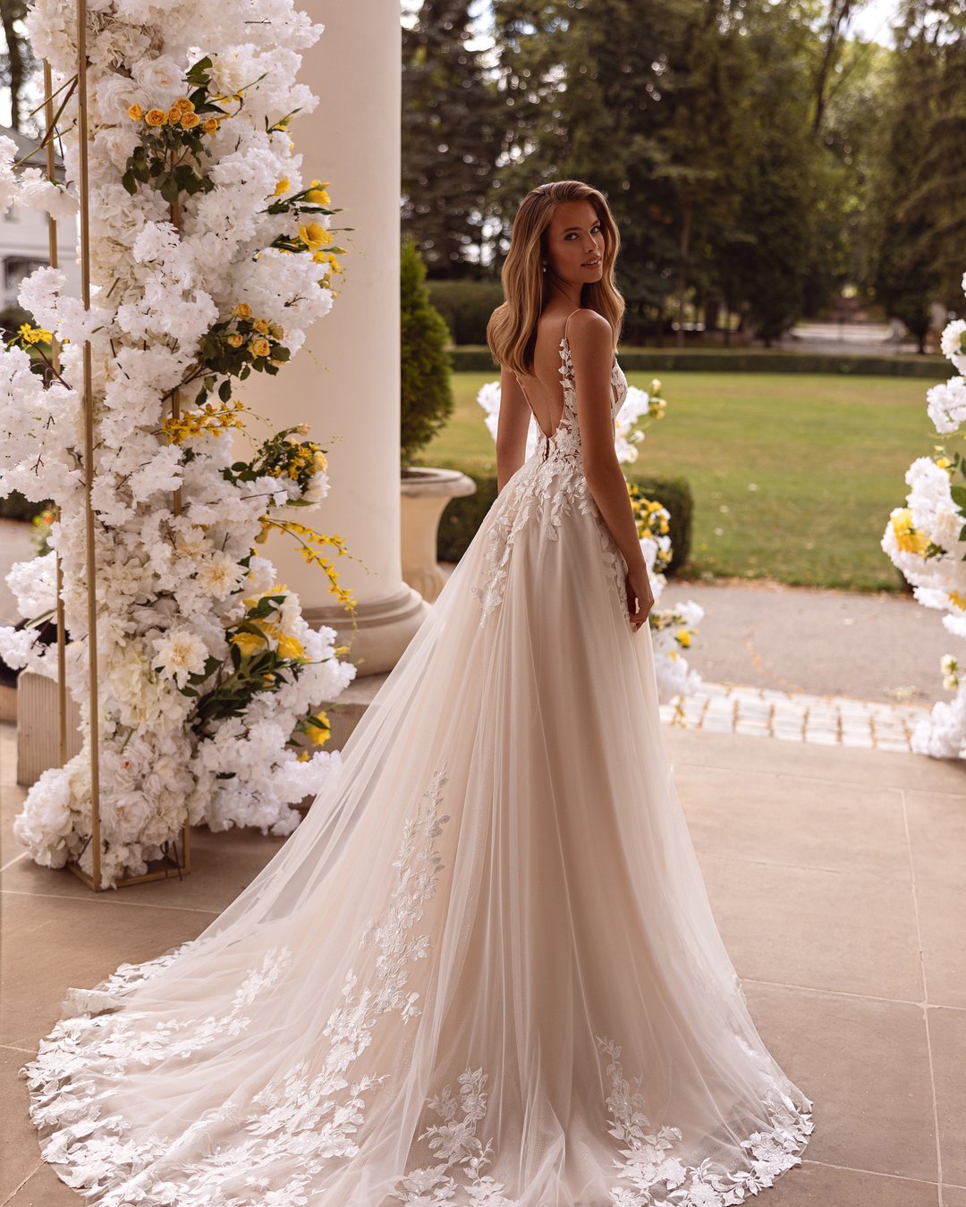 Suknia ślubna Kushni z kolekcji AnnaSposa to prawdziwa kwintesencja romantyzmu i elegancji. Odkryte plecy oraz szeroka spódnica w linii A, ozdobiona delikatną koronką i tiulem, sprawiają, że każda panna młoda poczuje się jak w bajce. Efektowny tren i subtelne ramiączka podkreślają wyjątkowość tej kreacji, a jej subtelny blask doda magii każdemu ślubnemu momentowi.