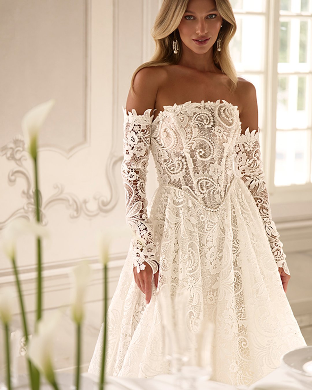 Suknia ślubna Bohnia od Anna Sposa to elegancka princeska z szeroką spódnicą i transparentnym gorsetem. Wykonana z bogato zdobionej gipiury, ozdobiona perłami, z prostym dekoltem i odpinanymi rękawami. Idealna dla panny młodej marzącej o bajkowym wyglądzie.