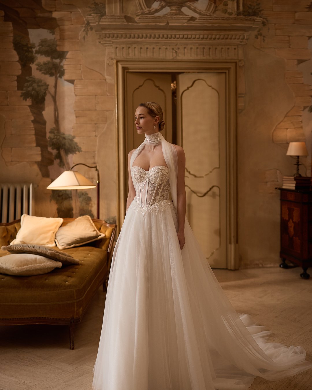 Suknia ślubna 5806 z kolekcji Elizabeth Passion to prawdziwe dzieło sztuki, które łączy nowoczesny styl z romantycznym wdziękiem. Szeroka spódnica z tiulu i koronki, ozdobiona subtelnymi kwiatowymi detalami, w połączeniu z gorsetową górą i eleganckim chokerem, podkreśla sylwetkę, a długi tren dodaje majestatyczności. Ta niezwykła suknia z pasem w kształcie litery V wprowadzi każdą pannę młodą w bajkowy świat miłości i elegancji.