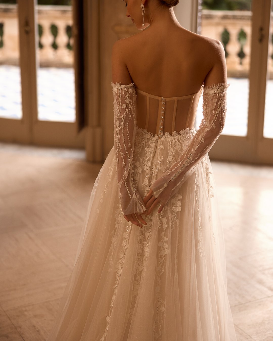 Suknia ślubna 5831 z kolekcji Elizabeth Passion łączy nowoczesny design z romantycznym wyrafinowaniem. Przezroczysta góra ozdobiona delikatną koronką i kwiatowymi aplikacjami, w połączeniu z szeroką, tiulową spódnicą, tworzy niepowtarzalny efekt, który zachwyca blaskiem. Nakładane rękawy dodają całości subtelności, sprawiając, że każda panna młoda poczuje się jak prawdziwa królewna w tym wyjątkowym dniu.