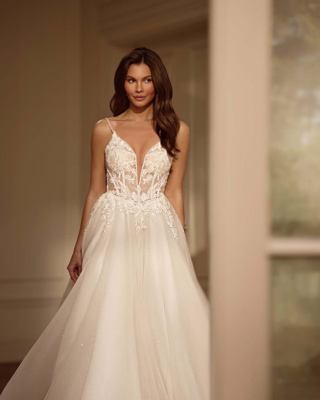 Zjawiskowa suknia w linii A na cienkich, delikatnych ramiączkach. Góra sukni wykonana z transparentnego tiulu zdobionego ręcznie wyszywaną koronką. Głęboki dekolt w kształcie litery V subtelnie eksponuje sylwetkę, a pięknie wycięte, koronkowe plecy dodają sukni romantyzmu i lekkości.