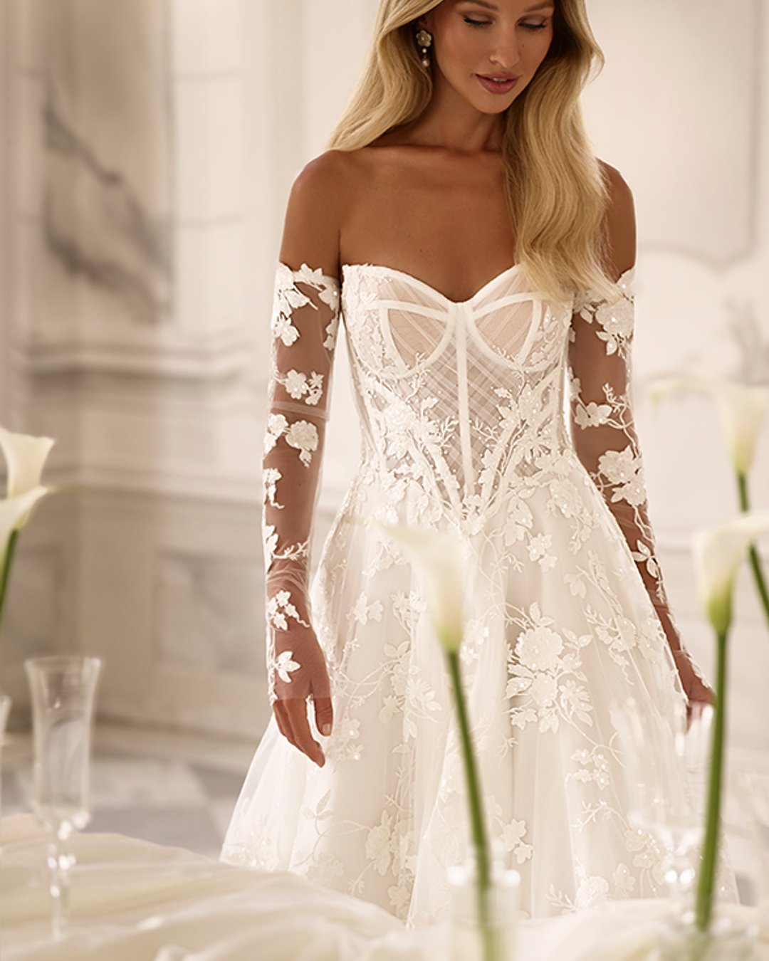 Subtelna suknia Reinbow zachwyca lekkością, romantycznymi kwiatowymi zdobieniami i nowoczesnym krojem w linii A. Gorsetowa góra, przezroczyste detale oraz opcjonalne rękawy tworzą stylową i kobiecą całość.