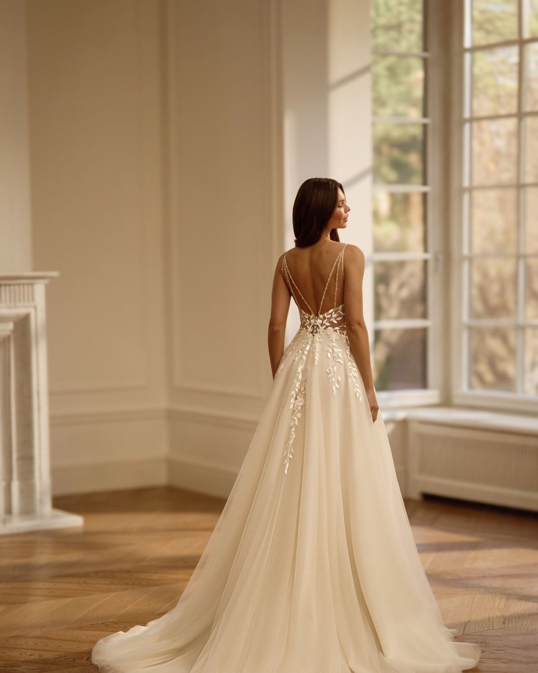 Suknia wykonana z tiulu na cienkich ramiączkach z wyciętymi, przezroczystymi bokami, które pięknie modelują sylwetkę.
Gorset zdobiony koronką w kształcie listków, głęboki dekolt w kształcie litery V oraz efektownie wycięte plecy na cienkich, ozdobnych ramiączkach.
Szeroka, lekka spódnica z rozcięciem na nodze dodaje subtelnego seksapilu.
Przezroczysta góra, lekki tiulowy tren.