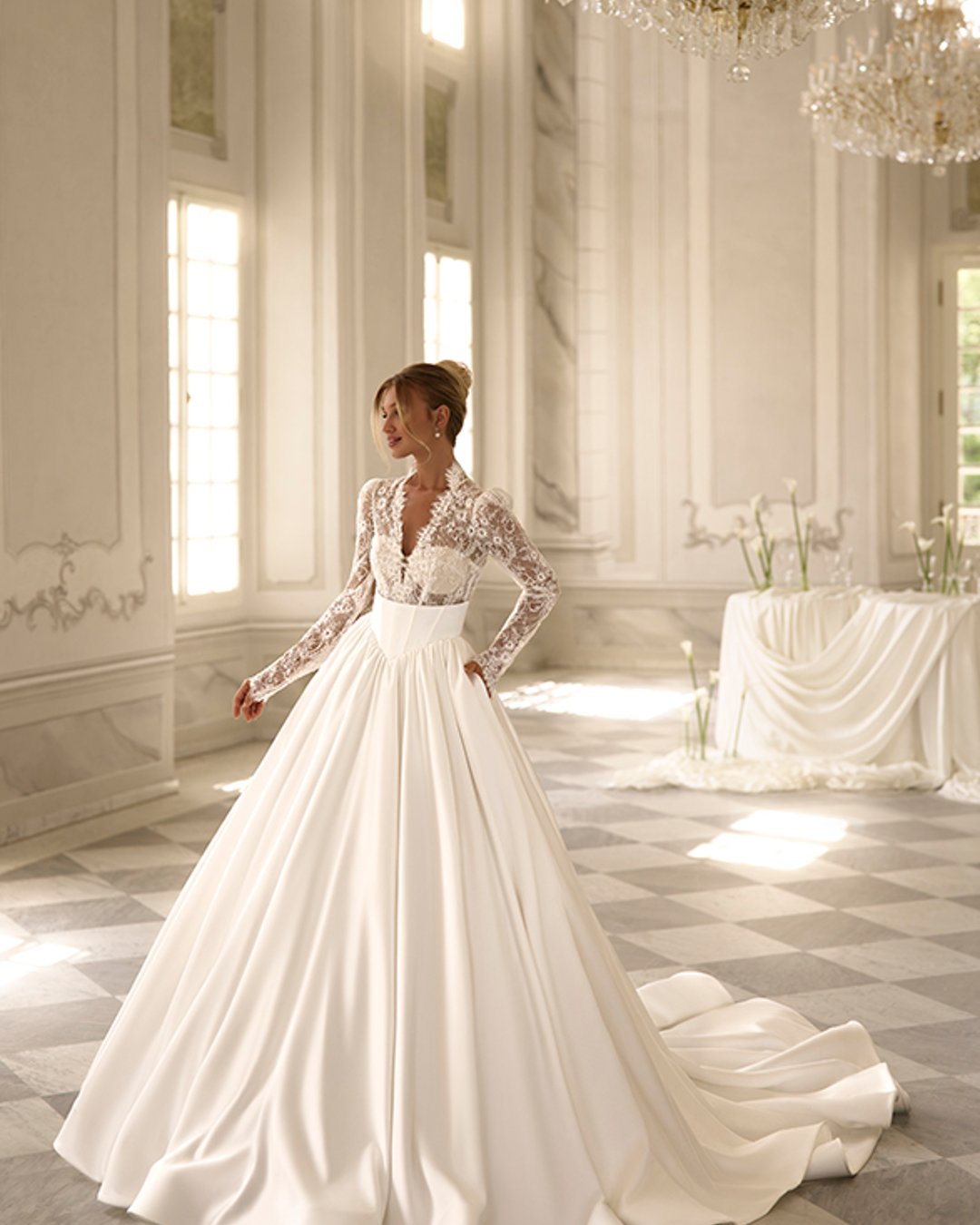 Calayra od Anna Sposa to elegancka suknia ślubna typu princessa, która zachwyca zmysłowym dekoltem w kształcie litery V, długimi koronkowymi rękawami oraz olśniewającą, rozkloszowaną satynową spódnicą z trenem. To połączenie klasyki i nowoczesnej delikatności.