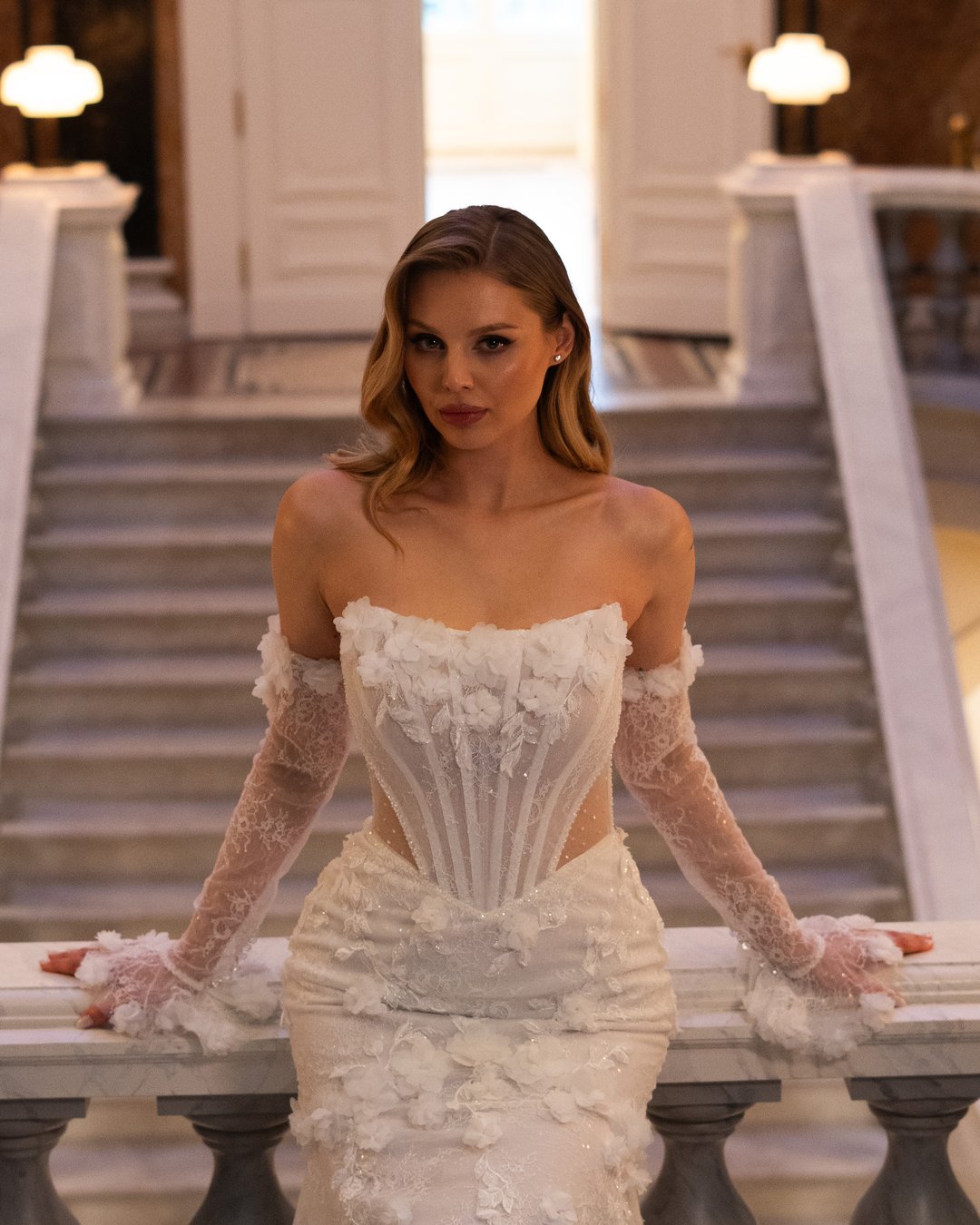 Suknia ślubna Charlotte z kolekcji LaLiven to prawdziwa ucieleśnienie elegancji i nowoczesnego romantyzmu. Jej koronkowy gorset z efektownym pasem w kształcie litery V oraz nakładane rękawy doskonale podkreślają sylwetkę, a błyszczące detale i kwiatowe motywy dodają subtelnej finezji. Model w stylu syrenki, idealnie dopasowany, sprawi, że każda panna młoda poczuje się wyjątkowo w swoim wielkim dniu.