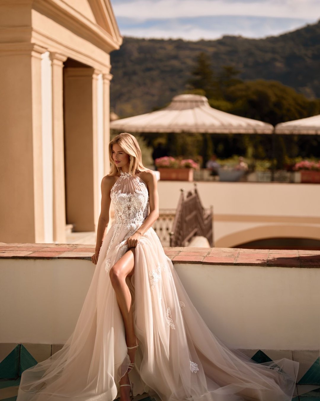 Iltana to romantyczna suknia ślubna z dekoltem halter, wyciętymi plecami i dekoracyjnymi aplikacjami w kształcie liści. Zachwyca szeroką, tiulową spódnicą, delikatnym połyskiem i subtelnym trenem.