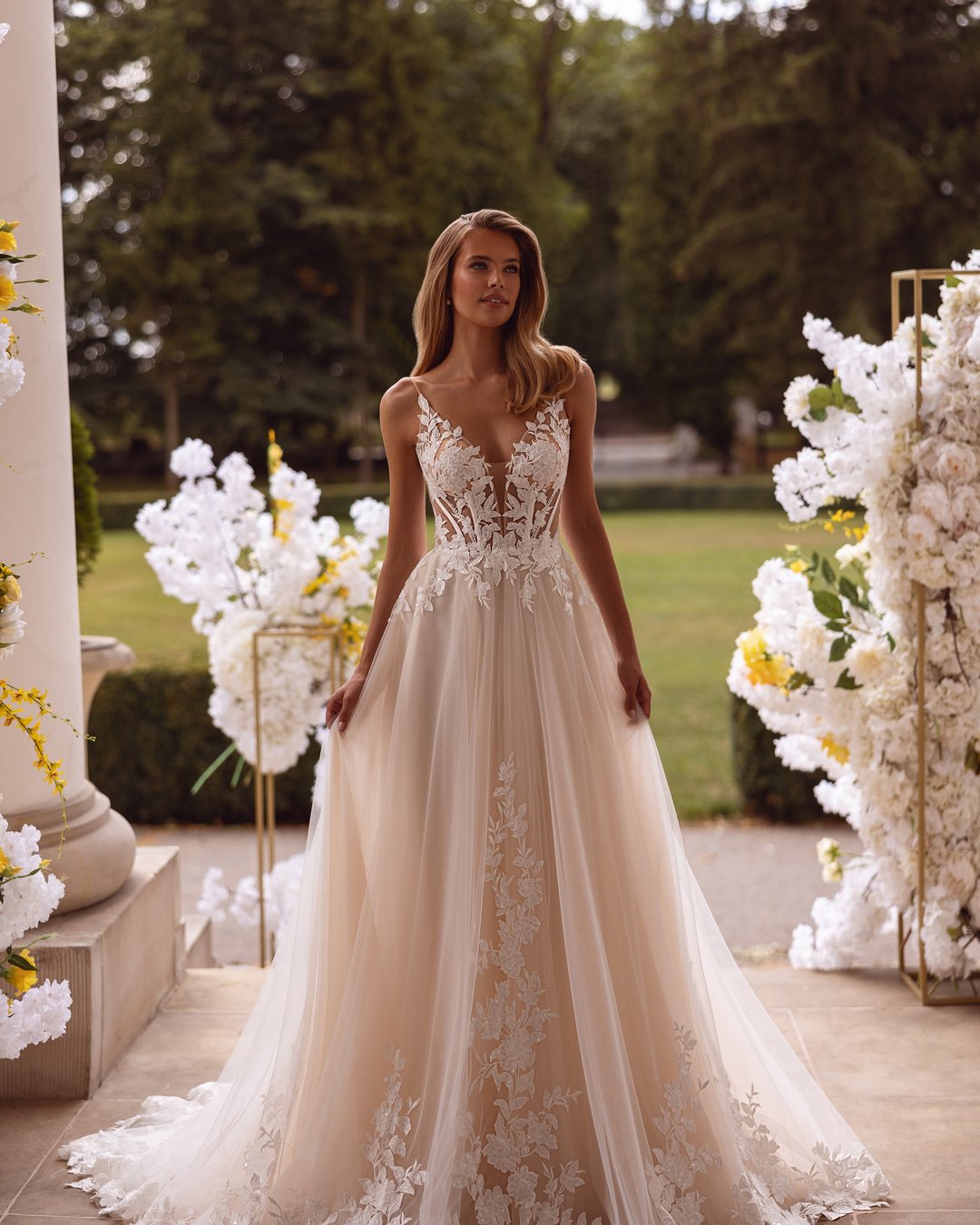 Suknia ślubna Kushni z kolekcji AnnaSposa to prawdziwa kwintesencja romantyzmu i elegancji. Odkryte plecy oraz szeroka spódnica w linii A, ozdobiona delikatną koronką i tiulem, sprawiają, że każda panna młoda poczuje się jak w bajce. Efektowny tren i subtelne ramiączka podkreślają wyjątkowość tej kreacji, a jej subtelny blask doda magii każdemu ślubnemu momentowi.