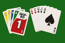 A high-angle, eye-level shot shows two stacks of cards on a green felt. On the left is a colorful stack of Skip-Bo cards. The top card is red with a large white "1." Underneath are a green "4," a blue card, and a yellow card. The stack on the right is a fan of five playing cards from a standard deck. The cards are all spades and are fanned out in ascending order, from the Jack to the Ace. The cards are clean and new, with the white space around the suits and numbers being bright and crisp. How to play Skip-Bo with regular cards.