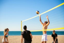Group playing beach volleyball on a sunny day with blue sky and ocean in the background.