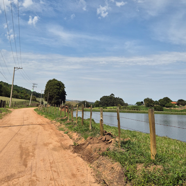 Estrada de terra margeando cerca de arame e um lago sob céu parcialmente nublado