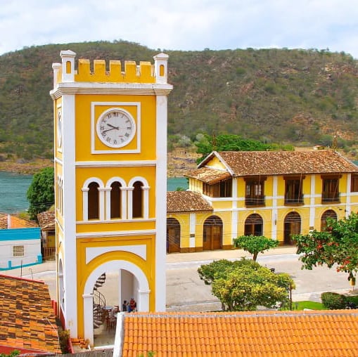 Torre amarela com relógio e casarão colonial às margens de um lago cercado por montanhas