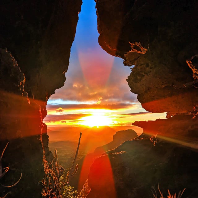 Pôr do sol visto através de uma abertura entre rochas, com raios de luz e vale ao fundo