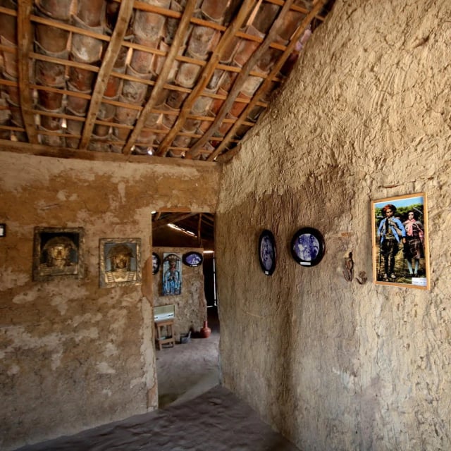 Interior de casa de taipa com teto de madeira e telhas, com quadros e fotos antigas nas paredes