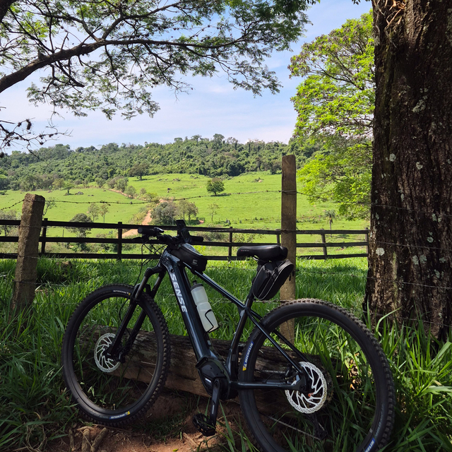 Bicicleta de trilha encostada em cerca de madeira, com pasto verde e morros ao fundo