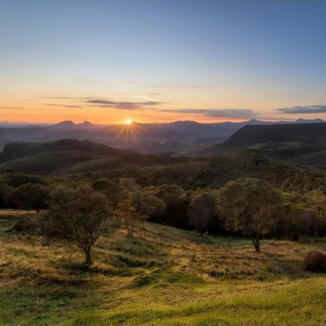 Sol nascendo entre serras e campos com árvores em uma paisagem de montanha ao amanhecer