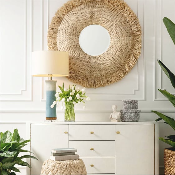 Large round raffia mirror hung over a white console table styled with a blue table lamp and white tulips.