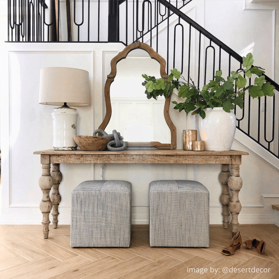 Inviting entryway featuring a reclaimed wood console table adorned with a mirror, candles, and a lamp. A ceramic vase with lush greenery sits gracefully, set against the backdrop of a captivating black wrought iron staircase.