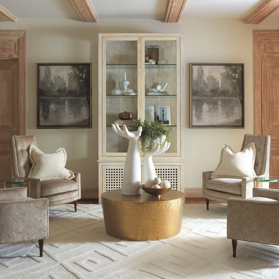 Sitting area with a gold hammered finished coffee table, adorned by two large hand-shaped candle holders. Four matching beige upholstered chairs encircle the table, set against a backdrop of a bookcase with artwork hung on each side, creating a harmonious and inviting arrangement.