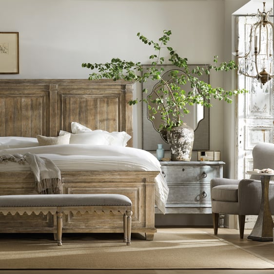 A bright bedroom with a French Country style wooden headboard, antique style nightstand with a weathered vase full of leafy branches, and a sitting area in front of a window with a chandelier hanging above an armchair.