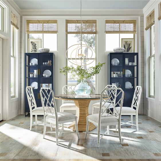 Bright dining room featuring a round rattan table with a glass top, encircled by white bamboo chairs. Adjacent to the table, two matching blue cabinets add a touch of color. The room is illuminated by numerous windows adorned with matchstick roller shades.
