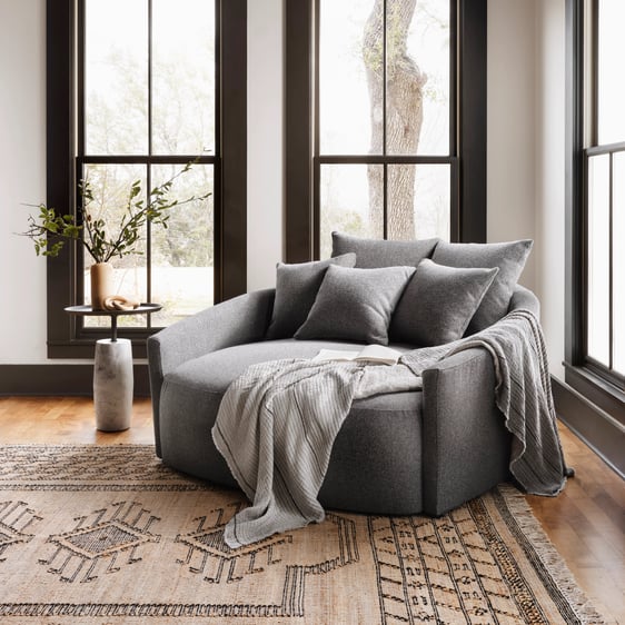Corner view of a spacious gray lounge chair adorned with stylish throw pillows and a cozy throw blanket. The chair is positioned in a well-lit room with expansive windows, allowing ample natural light to illuminate the scene. A side table rests beside the chair, and a patterned rug lies underneath, adding warmth and texture to the setting.