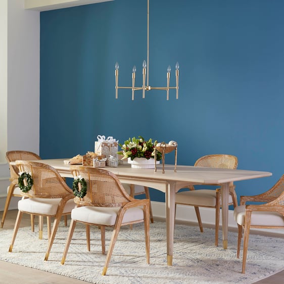 Dining room with blue accent wall featuring a dining table and dining chairs with miniature wreaths hanging over the back of the chairs. Wrapped present and accessories from Villa & House sit on top of the table.