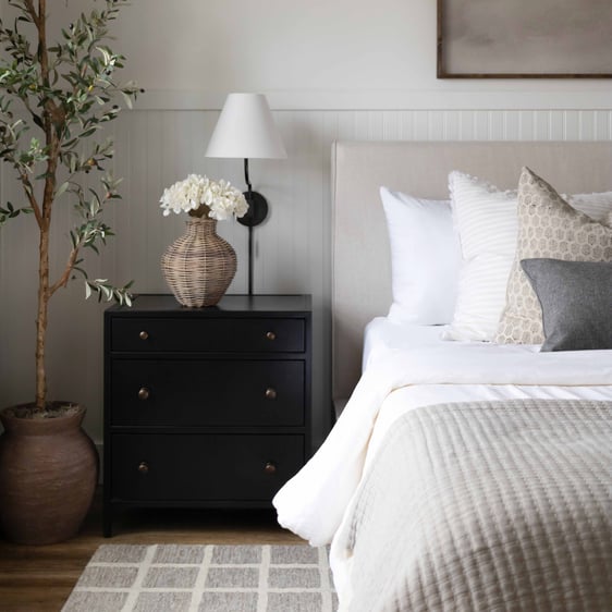 Black, three-drawer nightstand next to bed with neutral bedding. A black wall sconce hangs above the nightstand and a wicker vase with white flowers sits on top.