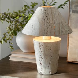 Modern travertine table lamp with tapered base and wide shade, styled on a wood console with books and ceramic accents.