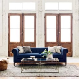A deep blue sofa is centered behind a thin glass coffee table with two large wooden double doors behind the furniture.