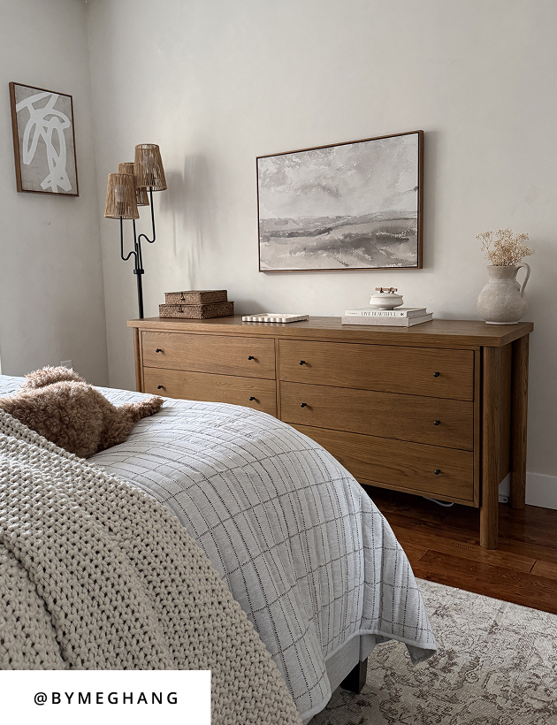 Warm neutral bedroom styled with wood dresser, layered bedding, and minimalist wall art