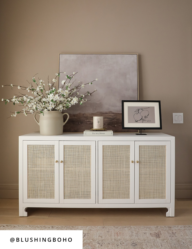 White cane cabinet with neutral art, floral arrangement, and minimalist decor on beige wall.