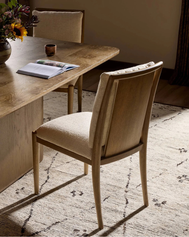 Upholstered dining chair at wood table on neutral patterned rug with warm lighting.