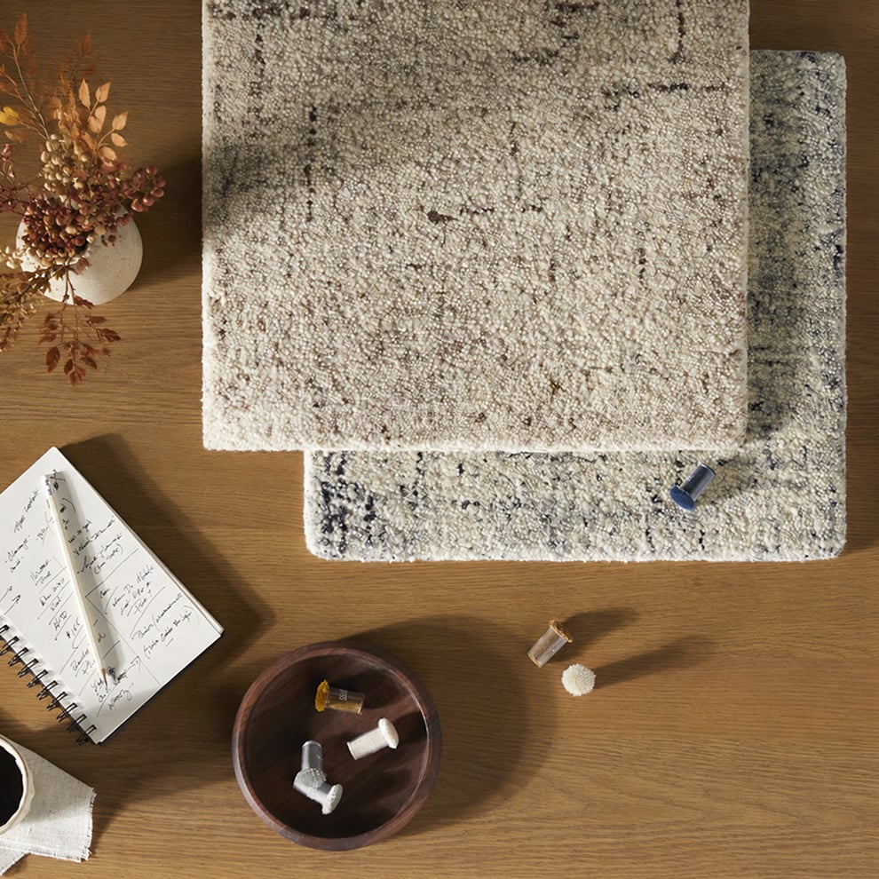 Top view of textured rug swatches on wood table with sewing tools and notebook.