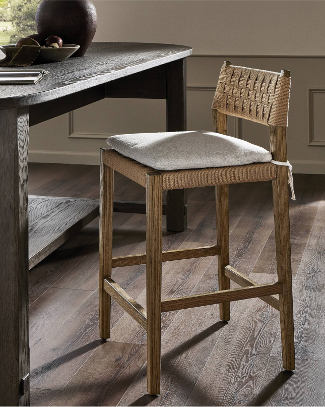Woven wood bar stool with cushion at dark wood table on hardwood floor.