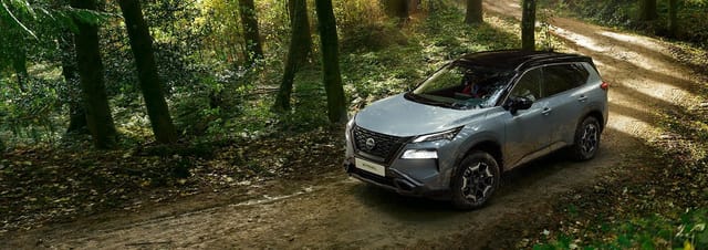 Een grijze Nissan X-Trail SUV op een onverharde weg in een dicht bos met bomen en schaduw