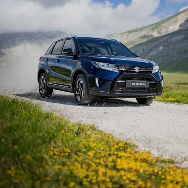 Een blauwe Suzuki Vitara SUV rijdend op een grindweg in een bergachtig gebied, omgeven door gras en gele bloemen onder een deels bewolkte hemel.