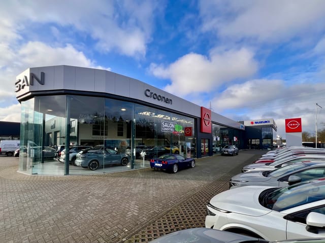 Moderne autodealer met Nissan en Suzuki merken, glazen gevel en geparkeerde auto's buiten onder een blauwe lucht met wolken