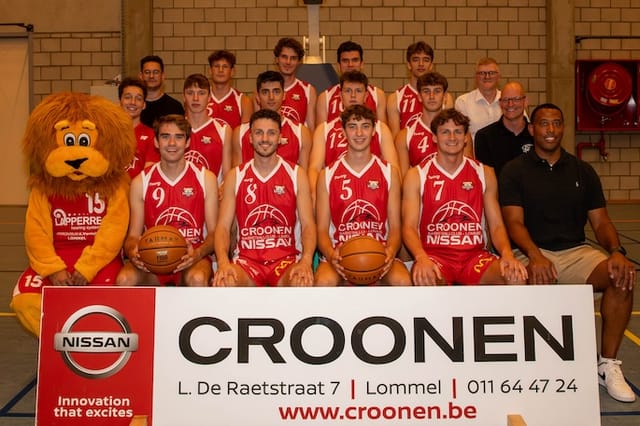 Basketbalteam groepsfoto met spelers in rode jerseys, mascotte in leeuwkostuum en coaches, voor een Croonen Nissan sponsorbord.