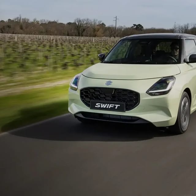 Een lichtgroene Suzuki Swift auto rijdend op een weg met groen en bomen op de achtergrond