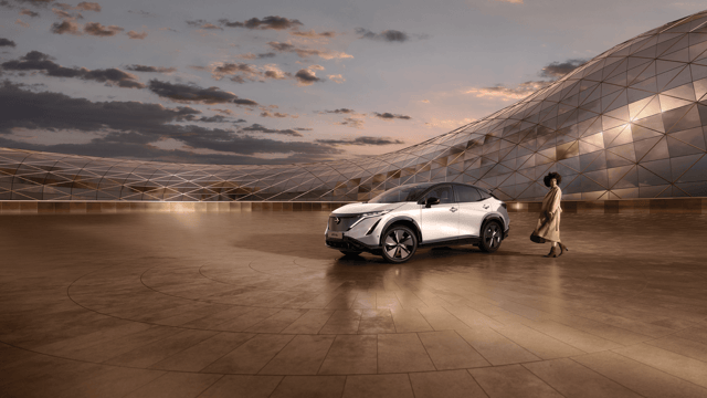 Moderne zilverwitte Nissan Ariya elektrische auto geparkeerd voor futuristisch gebouw met glazen gevel, vrouw in beige jas loopt ernaast