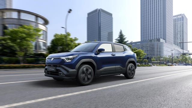 Een blauwe Suzuki Vitara SUV die over een stadsweg rijdt met moderne hoogbouw en groen op de achtergrond.