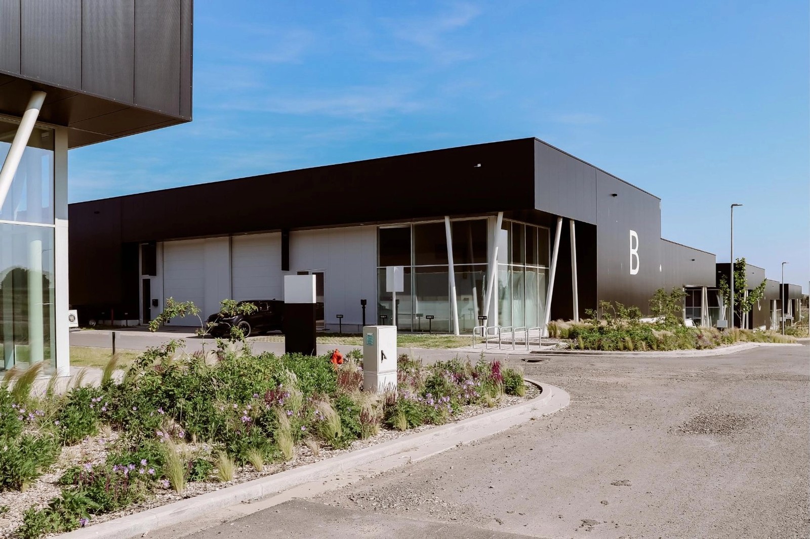 Complexe de bâtiments industriels modernes avec façade noire et grandes fenêtres en verre, entouré de végétation et d'une voie pavée.