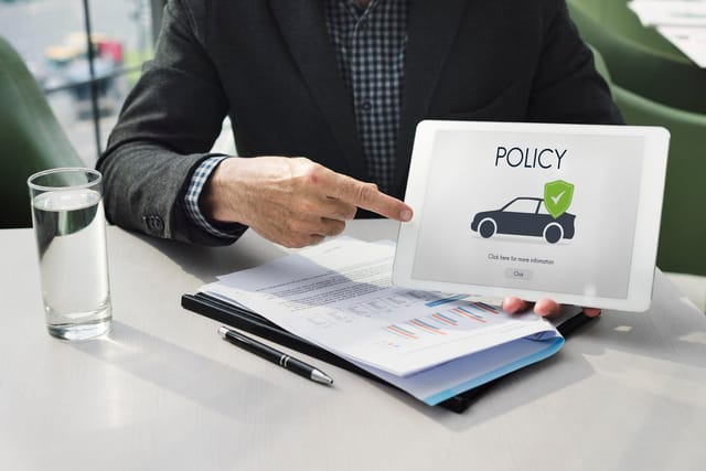 Iemand in zakelijke kleding wijst naar een tablet met een afbeelding van een auto en het woord 'POLICY', op een bureau met documenten en een glas water.