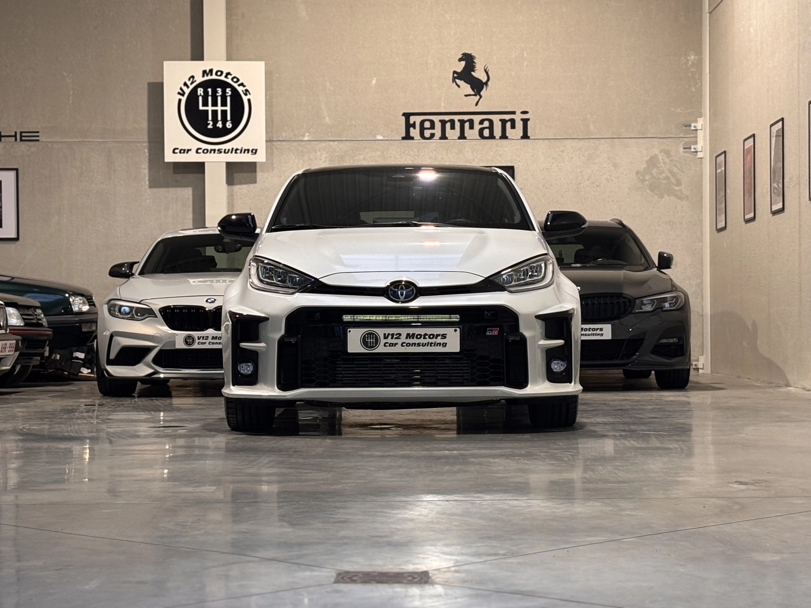 Une voiture Toyota blanche en premier plan dans un espace intérieur, avec derrière une BMW blanche et une BMW noire, toutes portant des plaques V12 Motors.