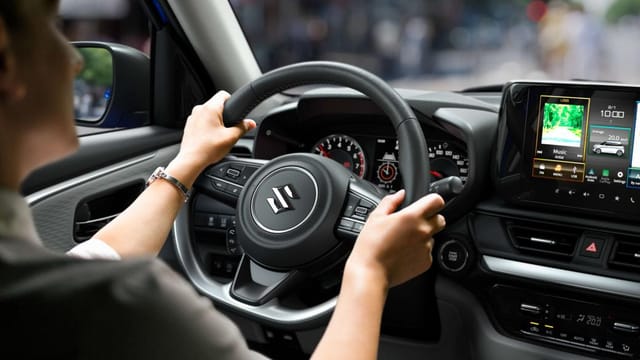 Persoon houdt het stuur van een Suzuki-auto vast met het dashboard en het infotainmentscherm zichtbaar.