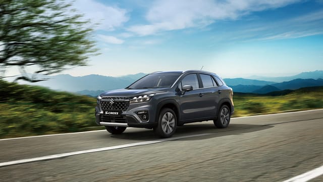 Donkergrijze Suzuki S-Cross SUV rijdend op een open weg met een vervaagde boom links en berglandschap op de achtergrond onder een deels bewolkte blauwe hemel.