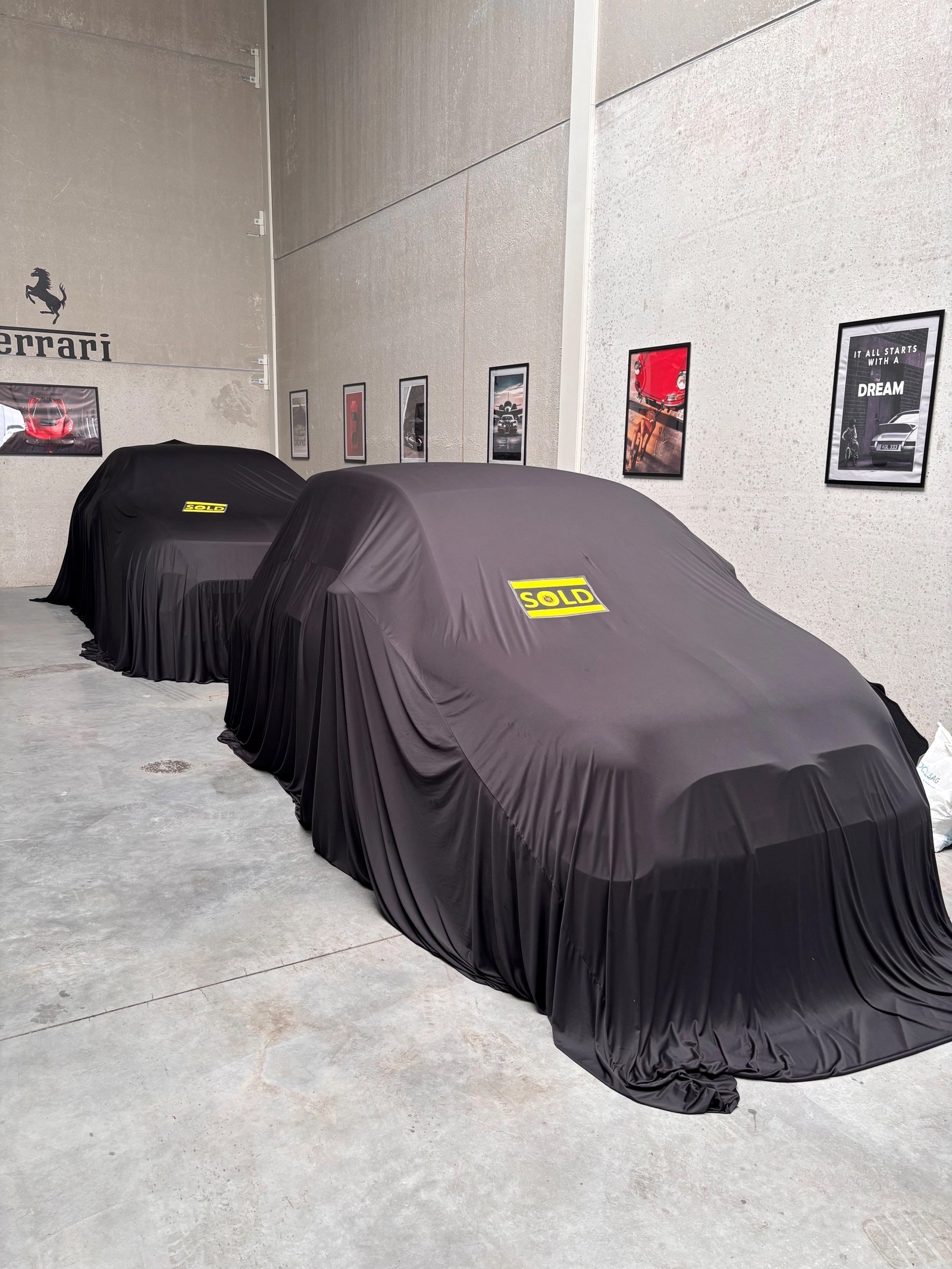 Deux voitures couvertes de draps noirs dans une salle d'exposition, toutes deux avec des panneaux jaunes "VENDU".
