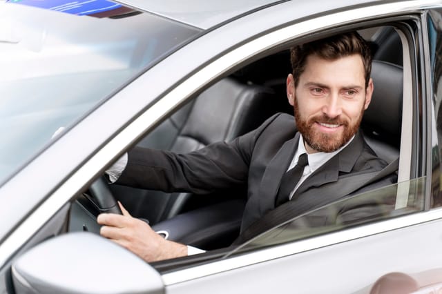Een glimlachende man met baard in een zwarte jas rijdt een auto en kijkt uit het raam