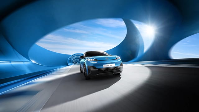 Een blauwe Nissan Micra die door een futuristische tunnel met grote ovale ramen rijdt, met zonlicht aan de zijkant